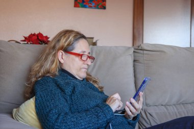 Olgun bir kadın koltukta dinlenirken cep telefonunu kullanıyor.