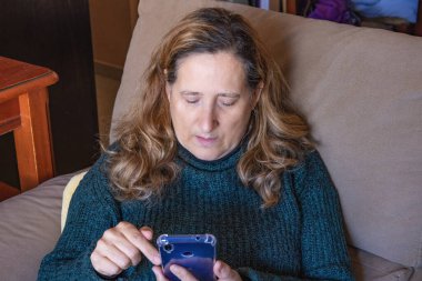 Olgun bir kadın koltukta dinlenirken cep telefonunu kullanıyor.