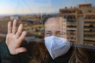 Coronavirus Covid-19 tarafından izole edilmiş bir kadın koruyucu bir maske takarak evinin camından bakıyor.