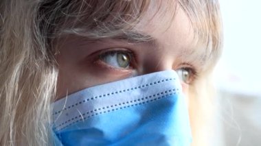 Coronavirus Covid-19 tarafından izole edilmiş genç bir kadın koruyucu bir maske takarak evinin camından bakıyor.
