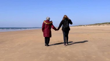 Olgun bir kadın, yaşlı annesiyle sonbaharda kumsalda yürüyor.