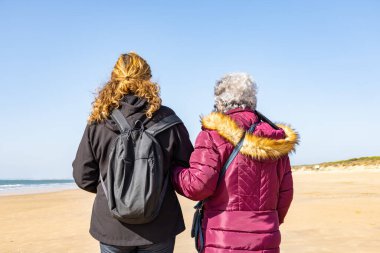 Olgun bir kadın, yaşlı annesiyle sonbaharda kumsalda yürüyor.