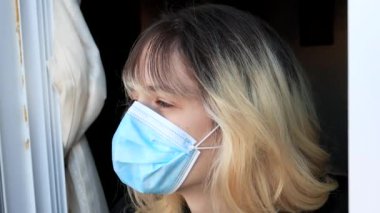 Coronavirus Covid-19 tarafından izole edilmiş genç bir kadın evinin camından dışarı bakıyor ve koruyucu bir maske takıyor.