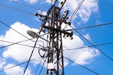 High voltage electricity pole, street lamp and blue sky with white clouds.