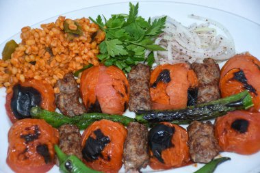 Domates kebabı, bulgur pilavı, kavrulmuş biber, patlıcan ve soğanlı maydanozla servis edilir.. 