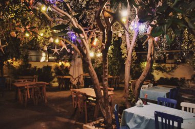 Bahçesinde masalar olan tipik Yunan tavernası. Ege kasabasındaki bir kır bahçesinde restoran. Lambalar, masalar ve renkli ağaçlardan sarkan sandalyeler. Yunan tavernası