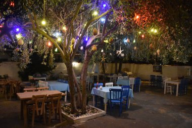 Bahçesinde masalar olan tipik Yunan tavernası. Ege kasabasındaki bir kır bahçesinde restoran. Lambalar, masalar ve renkli ağaçlardan sarkan sandalyeler. Yunan tavernası
