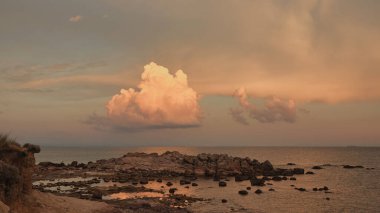 Gökyüzünde kırmızı, beyaz ve kıpkırmızı tonlarda asılı büyük bulut kütleleri ve kumsalda kırmızı kayalıklar ve çakıl taşları
