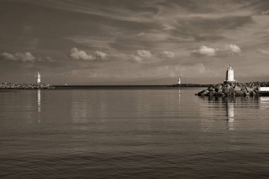Limanın köşesinde duran eski bir deniz feneri. Dalgalar sorunsuzca sallanıyor. Mavi açık gökyüzü, deniz üzerinde beyaz sirrus bulutları. Turuncu, sarı renkler baskın. Bw vintage.