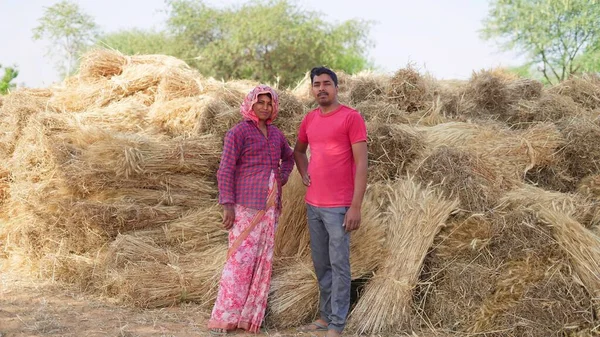 Young Indian farmer standing with his wife in front of wheat crop pile.