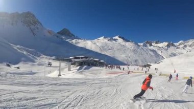 Turuncu takım elbiseli bir snowboardcu yamaçtan aşağı iniyor.