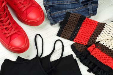 Yakın plan, kadınlar için günlük kıyafet ayrıntıları - mavi kot, kırmızı spor ayakkabı, siyah kolsuz kolsuz bluz ve çok renkli, beyaz ahşap arka planda elle örülmüş pamuk bluz