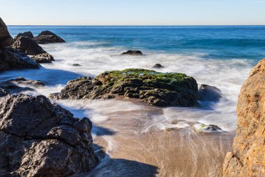 Malibu, California 'daki Rocky plajı. Dalgalar kumlu sahilden çekiliyor; mavi Pasifik Okyanusu arka planda. 