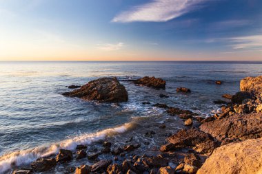 Sabahın erken saatlerinde Malibu, Kaliforniya 'daki Rocky plajı. Uzakta sakin Pasifik Okyanusu; mavi gökyüzü ve üzerinde bulutlar.