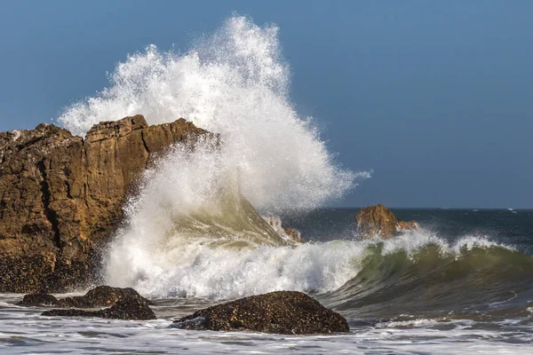 Malibu, California yakınlarında dev kaya oluşumuna karşı büyük dalga kırılıyor. Sprey havada yüksekten uçuyor. Gökyüzü, uzaktaki Pasifik Okyanusu. 
