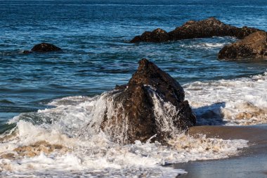 Malibu, California yakınlarındaki bir sahilde, gelgitte açığa çıkan bir kayadan su çekiliyor. Arka planda Mavi Pasifik Okyanusu.