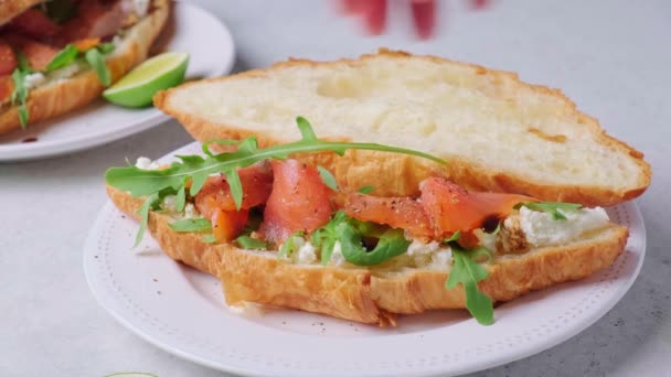 Sandwich croissant avec fromage à la crème, saumon et roquette sur assiette blanche. Concept de petit déjeuner sain. — Video