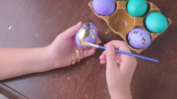Dłonie dzieci zdobią pisanki w złotym liściu przy drewnianym stole w domu. — Wideo stockowe