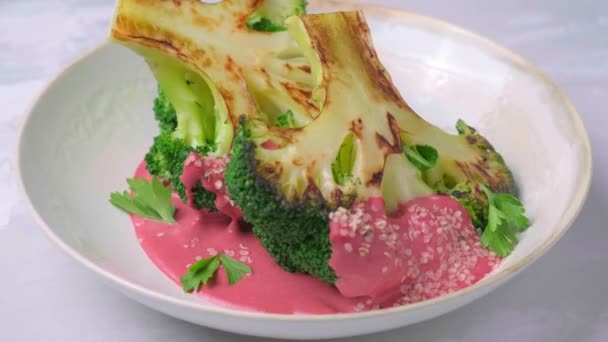 Gegrilltes Brokkoli-Steak mit rosa Rote-Bete-Sauce und Hanfsamen in weißer Schüssel, Teller dreht sich. — Stockvideo