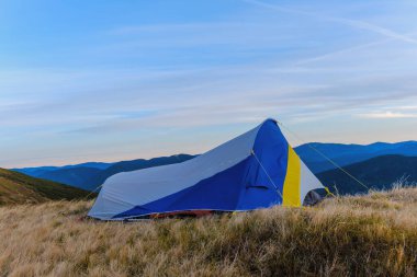 Sarı otlarla kaplı bir yamaçta dağ çadırı. Arka planda, dağ sıraları ve mavi gökyüzü.