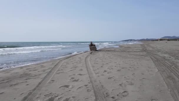 Drone avec caméra se déplace rapidement vers l'homme et décolle sur l'eau. Guy est assis sur une chaise, se rafraîchissant et se prélassant au bord de la mer Adriatique. Des lignes sont tracées sur le rivage sablonneux autour de lui — Video