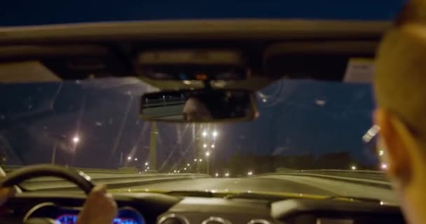 Mujer morena joven está conduciendo elegante coche convertible a través de la ciudad de la noche, y el chico de moda con pendiente en la oreja está sentado en el asiento del pasajero, la carretera es visible a través del parabrisas, vista desde el asiento trasero — Vídeos de Stock