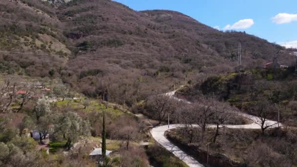 Drón kamerával emelkedik át dombok borított erdő, és az út fut keresztül egy falun a vidéken, gyönyörű kék ég mögött hegy a horizonton, panorámás kilátás — Stock videók