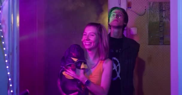 Adolescentes con estilo con cachorro dachshund en chaqueta de cuero vinieron a la fiesta de la casa, mirando a su alrededor. Apartamento está decorado con guirnaldas brillantes e iluminado con luz mística multicolor — Vídeos de Stock