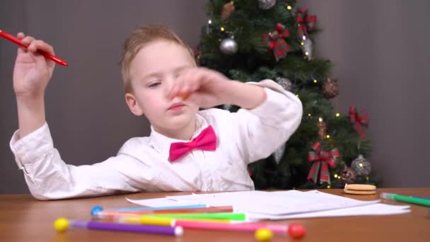 Мальчик с маркером ест конфеты за столом возле рождественской елки — стоковое видео