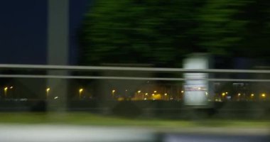 Araba gece kasabasının yolundan hızla geçer, sokak lambalarıyla aydınlatılmış caddeleri ve binaları geçerek, çitlerle çevrili şehir parklarını içerideki otomobillerin penceresinden görür. Diğer araçlar hızla geçiyor.