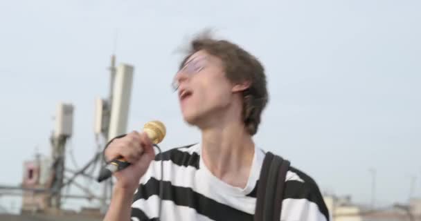 Молодий грабіжник в окулярах виразно співає з золотим мікрофоном і трясе голову на даху, його волосся розвіяне вітром. Відкритий сеанс варення — стокове відео