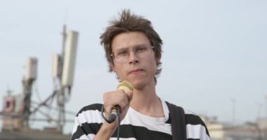 Gözlüklü ve burun piercingli genç adam mikrofonu standda tutuyor, elektro gitarla enerjik bir şekilde dans ediyor ve açık hava konserinde şarkı söylüyor.