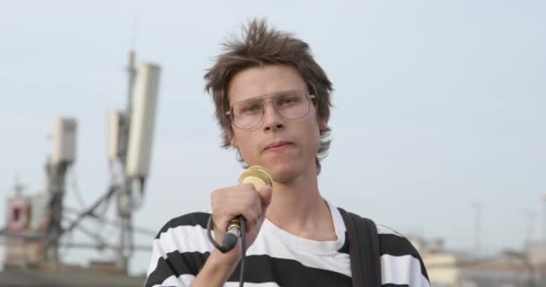 Joven con gafas y piercing en la nariz sostiene micrófono en un soporte, baila enérgicamente con la guitarra eléctrica y canta durante el concierto al aire libre, su tiene el pelo desaliñado con estilo — Vídeos de Stock