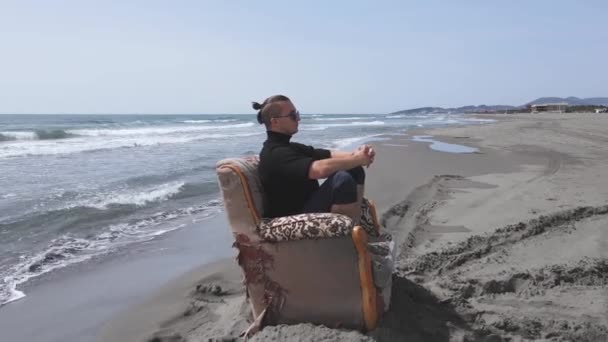 Беспилотник улетает от модного парня, сидящего в старом кресле в медитативной и вдумчивой позе, линии нарисованы на песчаном побережье в разных направлениях вокруг него, как лучи солнца, вид сбоку — стоковое видео