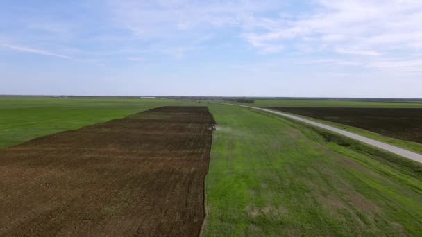 Панорамный вид сельскохозяйственных полей вдоль дороги, вдалеке тракторные плуги и бороны земли со специальным оборудованием, подготовка почвы для сезонного посева пшеницы, стрельба беспилотником с высоты — стоковое видео