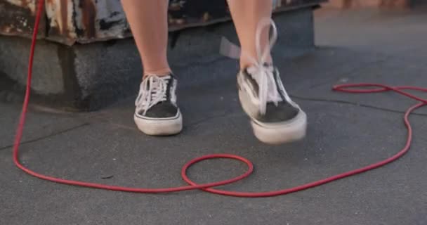 Osoba w czarno-białych tenisówkach skacze przez przewód elektryczny, stwarzając niebezpieczną sytuację, zamykając widok na nogi. Początkujący niezdarnie próbuje uprawiać sport i skakanie, skakanka — Wideo stockowe
