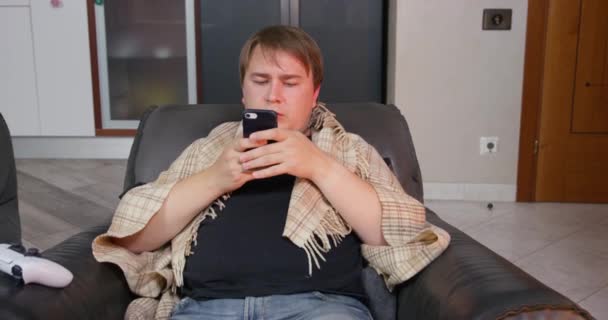 Jovem envolto em cobertor de lã está sentado em poltrona e entusiasticamente usando smartphone. Freelancer trabalha em casa. cara está constantemente jogando porque ele é viciado em jogos para celular — Vídeo de Stock