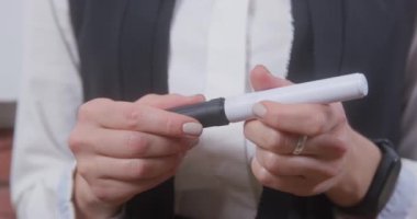 Beyaz bluzlu, düzgün manikürlü bir öğretmen sınıfta duruyor ve siyah keçeli kalemi son derece yakın çekimde açıyor.