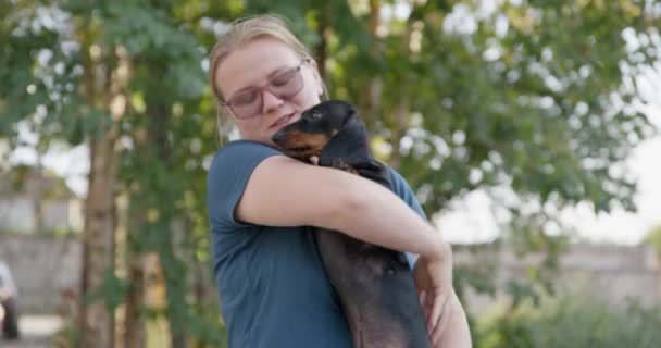 Jonge blonde vrouw met glazen knuffels en probeert een schattige teckel puppy te kussen, die het niet leuk vindt en probeert te ontsnappen aan de omhelzing tijdens een aangename wandeling in het park — Stockvideo