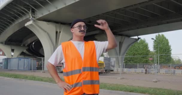 Trabajador con bigote gris grueso en visibilidad naranja y chaleco reflectante de seguridad inactivo en medio de la jornada laboral. Se para sin rumbo, mira a su alrededor y se rasca la nuca — Vídeo de stock