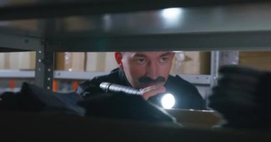Kalın bıyıklı ve siyah ceketli genç adam dikkatle rafları tarıyor. Karanlıkta, portatif bir el feneri parlıyor.
