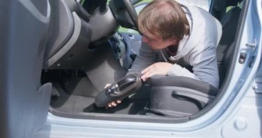 Şişko adam şoför koltuğunun altındaki arabayı elektrikli süpürgeyle temizliyor.