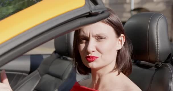 Junge brünette Frau mit spektakulärem Make-up und in roter Kleidung sitzt hinter dem Steuer eines Cabriolets und singt oder singt etwas, beleuchtet von hellen Sonnenstrahlen. — Stockvideo
