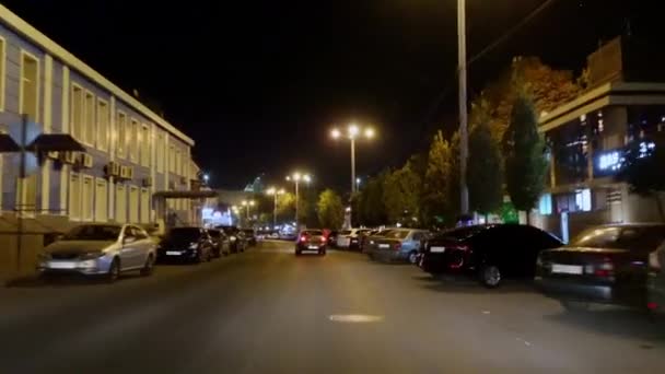 Bil kör längs smal gata i natten stad med restauranger, regeringskontor och parkeringsplatser, upplysta av lyktor, längs vilken människor går, skytte inifrån fordonet genom vindrutan — Stockvideo