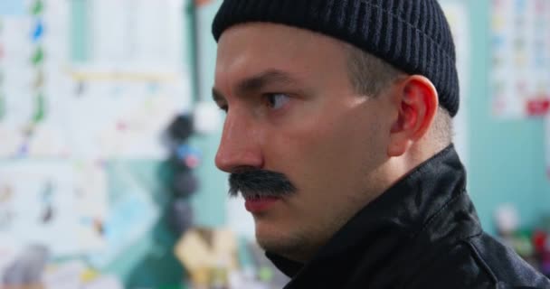 Young man with thick mustache in black knitted hat and jacket with collar turned up is watching something, when suddenly his attention is attracted by something from the side and he frowns — Stockvideo