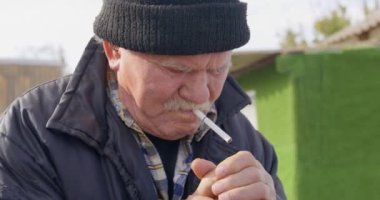 Gri kalın bıyıklı, kalın ceketli ve örgü şapkalı yaşlı bir adamın portresi. Sigarasını yakar ve bir fırt çeker, dışarı çıkar, ön tarafa bakar. Sağlıksız alışkanlıklar