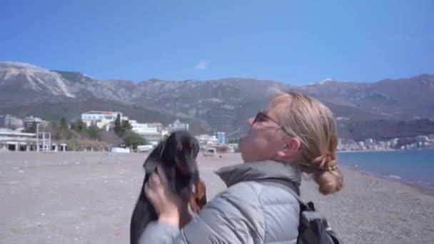Fiatal turista szőke nő sétál a tacskós kutyájával a tenger partján, feldobja a kiskutyát, megcsókolja. Nyaralás a tengerparton háziállatokkal — Stock videók