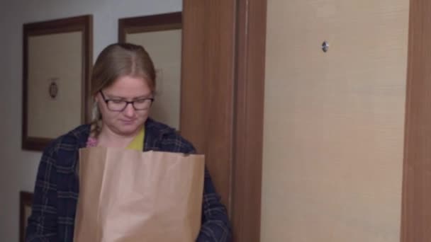 Pretty blonde woman with stylish glasses becomes scared and drops paper bag with products coming into light room, grabs his face with his hands — ストック動画