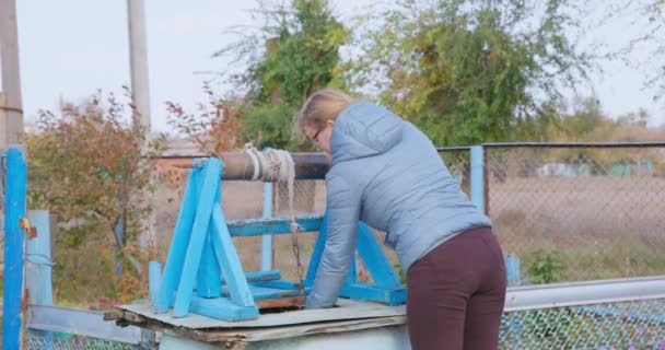 Aşağı ceketli genç sarışın kadın kuyuyu açar ve bir halata çinko kovayı indirir, temiz su toplamak için kulplu özel bir manuel mekanizma kullanır. Köyde günlük rutin — Stok video