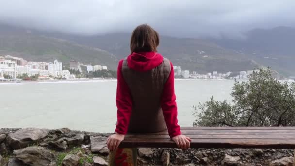 Женщина-туристка сидит на скамейке и смотрит на вид прибрежного города — стоковое видео